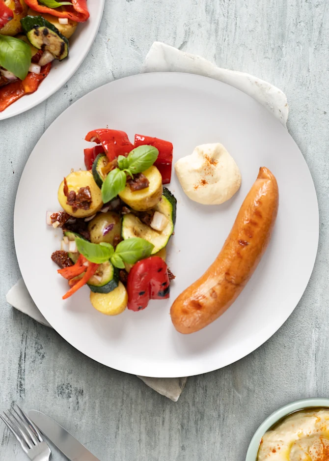 Grillpølser serveret med fyldig kartoffelsalat og grillede grøntsager. Hertil hummus